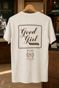 Load image into Gallery viewer, An off-white t-shirt hanging on a wooden hanger in a coffee shop setting, featuring a brown graphic print on the back with the text 'Good Girl Coffee Run' and 'Good Girl Beverages'.