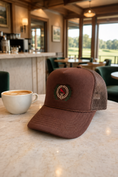 Load image into Gallery viewer, A brown trucker hat with a mesh back and a circular embroidered logo sits on a light marble table next to a white coffee cup in a cafe setting.