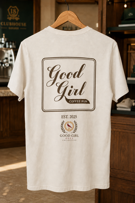 An off-white t-shirt hanging on a wooden hanger in a coffee shop setting, featuring a brown graphic print on the back with the text 'Good Girl Coffee Run' and 'Good Girl Beverages'.